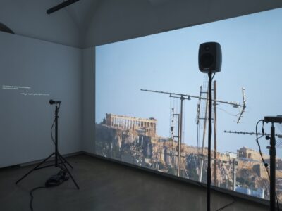 Foto Ausstellung mit Videos und Text an den Wänden, Lautsprecher und kleine Projektoren auf Stativen stehen am Boden