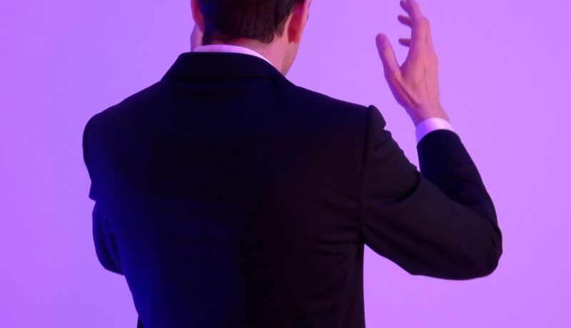 A person seen from behind, wearing a dark suit and a white shirt, with their hands raised against a purple background