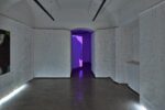 Photo of an exhibition featuring coloured pencil lines on white paper drawings on white paper rolls, and a video installation in the background