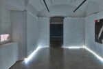 Photo of an exhibition featuring coloured pencil lines on white paper drawings on white paper rolls, and chalk drawings on a black wall in the background