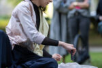 Documentation photo of the performance with a women sitting on a table in a garden and drips wax from a candle onto a piece of fabric surrounded by people