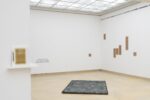 Photo of the exhibition at Kunstpavillon with a publication on a shelf leaning towards the wall, wooden panels and text on the wall and a fabric on the floor