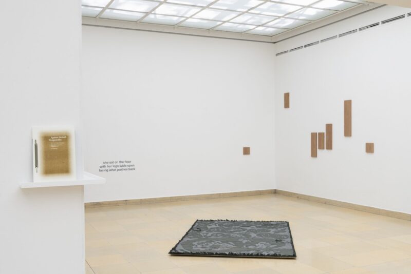 Photo of the exhibition at Kunstpavillon with a publication on a shelf leaning towards the wall, wooden panels and text on the wall and a fabric on the floor