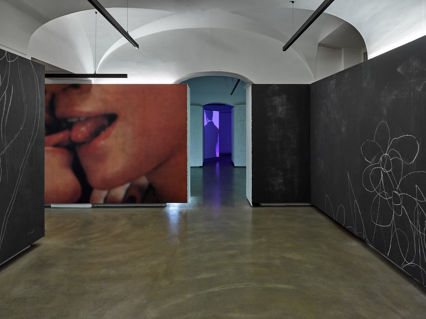 Foto Ausstellung mit Kreidezeichnungen auf schwarzer Wand, Foto von zwei sich berührenden Zungen und Videoinstallation im Hintergrund