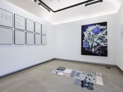 Foto Ausstellungraum mit gerahmten schwarz weiß Bildern links, Foto Blumenstrauß vor schwarzem Hintergrund und Assemblage am Boden