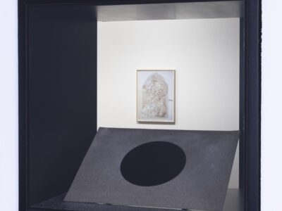 Ausstellungsansicht Blick durch Fenster in Galerieraum, glänzender Kreis auf beigem Filz und Sand, Bild an der Wand im Hintergrund